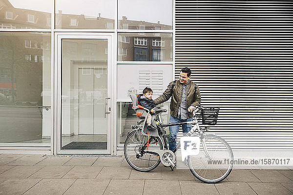 Lächelnder Vater fährt Fahrrad  während der Sohn im Sicherheitssitz auf dem Bürgersteig in der Stadt sitzt