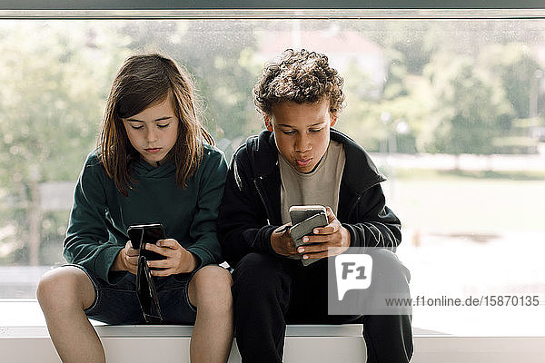 Schülerinnen und Schüler  die in der Schule vor einem Glasfenster sitzen und ein Mobiltelefon benutzen
