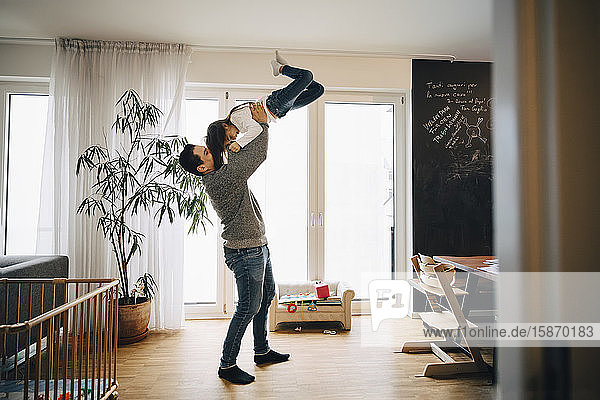 Verspielter Vater in voller Länge  der seine Tochter im Wohnzimmer stehend anhebt