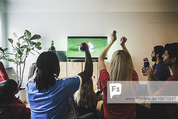 Fröhliche Freunde mit erhobenen Armen beim Fußballspiel im Wohnzimmer