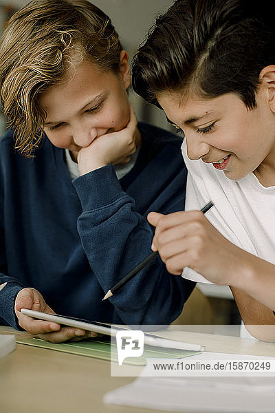 Männlicher Student zeigt auf digitales Tablett  während er mit einem Freund im Klassenzimmer sitzt