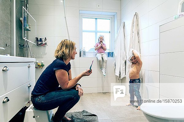 Seitenansicht der Mutter mit Blick auf die Tochter  die das Oberteil abnimmt  während das Mädchen am Fenster im Badezimmer sitzt
