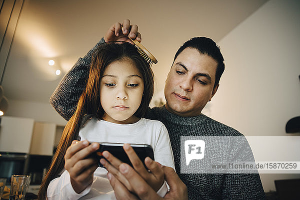 Niedriger Blickwinkel auf den Vater  der die Haare der Tochter kämmt  während das Mädchen zu Hause ein Mobiltelefon benutzt