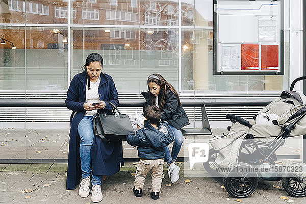 Mutter benutzt Smartphone  während sie mit Kindern an einer Bushaltestelle in der Stadt sitzt