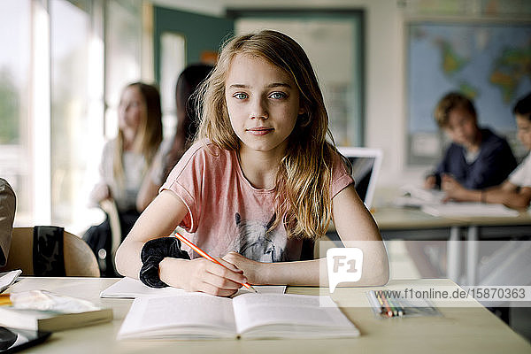 Portrait of female student writing in book while sitting at table in classroom