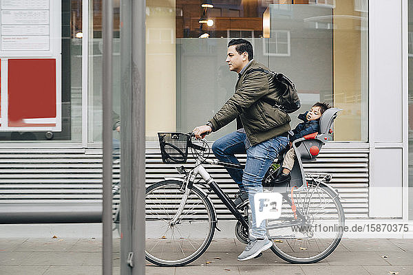 Vater fährt in voller Länge mit seinem Sohn Fahrrad auf dem Bürgersteig in der Stadt