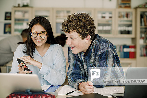 Smiling teenage girl showing smart phone to male friend while sitting in classroom