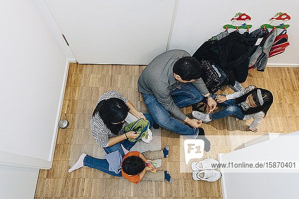 Schrägaufnahme eines Vaters  der der Tochter die Schnürsenkel bindet  während die Mutter mit dem Sohn zu Hause auf dem Boden sitzt
