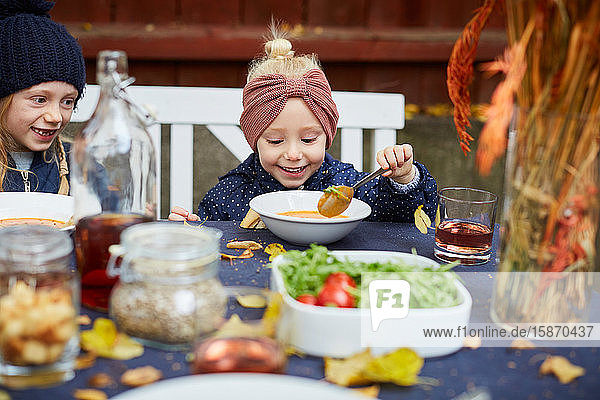 Lächelndes Mädchen isst vegetarische Suppe  während es neben einem weiblichen Geschwisterkind am Tisch sitzt