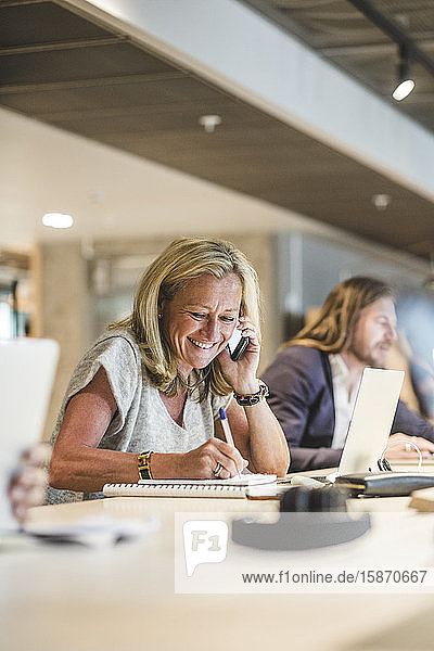 Lächelnde Geschäftsfrau schreibt Tagebuch  während sie im Büro am Smartphone telefoniert