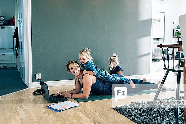 Lächelnde Mutter mit Laptop sieht Tochter auf dem Rücken liegend an  während das Mädchen zu Hause im Wohnzimmer sitzt