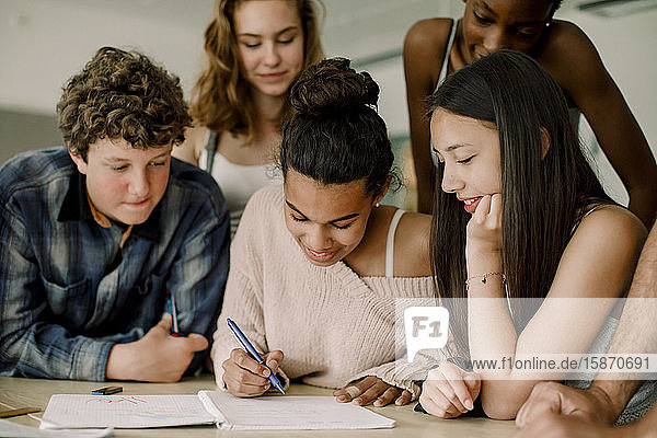 Lächelndes Teenagermädchen schreibt in Buch  während es inmitten von Freunden im Klassenzimmer sitzt