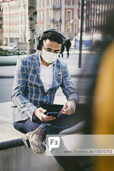 Geschäftsmann mit Smartphone  der draußen mit Gesichtsmaske sitzt