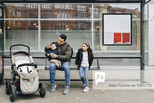 Vater gibt dem Sohn Babynahrung  während er mit der Tochter an der Bushaltestelle in der Stadt sitzt