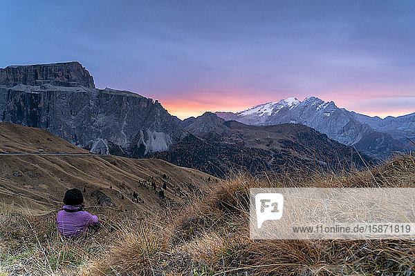 Rückansicht einer Frau  die im Gras sitzt und den Sonnenaufgang auf der Marmolada und dem Sass Pordoi  Sellajoch  Dolomiten  Südtirol  Italien  Europa  bewundert