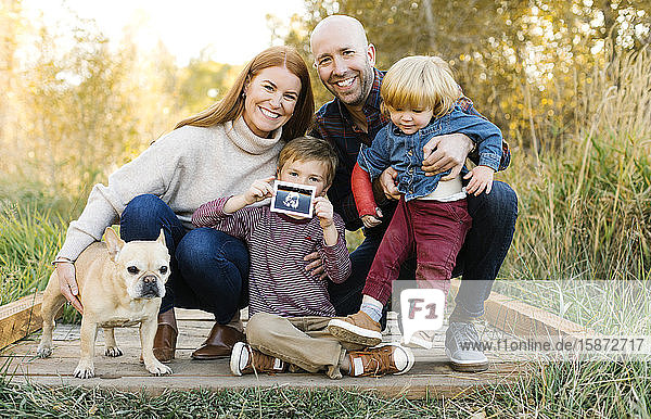 Smiling family with ultrasound photograph and pet dog on forest boardwalk