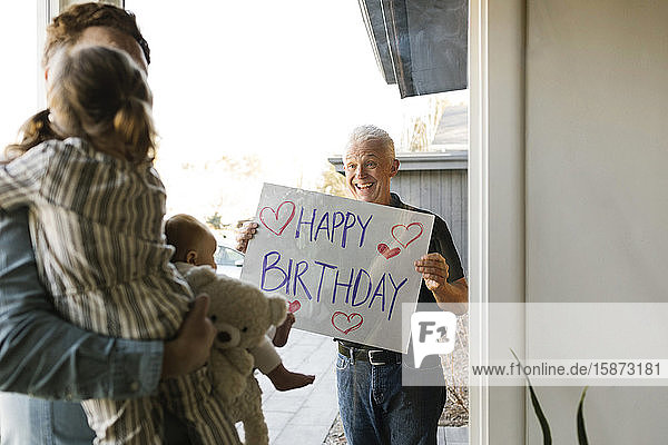 Großvater zeigt der Familie mit Enkelkindern (2-3 Monate  2-3) durch ein Fenster eine Glückwunschbotschaft zum Geburtstag