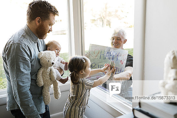 Großvater besucht Familie mit Enkelkindern (2-3 Monate  2-3) und zeigt Happy Birthday Zeichen durch das Fenster