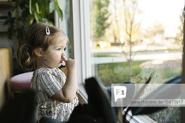 Girl (2-3) picking nose and looking through window