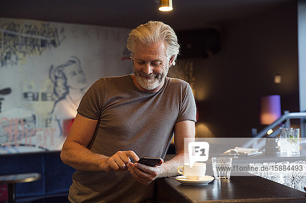 A man leaning on a bar beside a coffee cup  smiling and looking at a mobile phone.