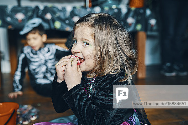 A girl smiling and eating sweets with a boy dressed as a skeleton in the background.