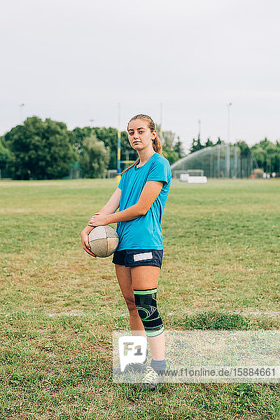Eine Frau steht auf einem Trainingsplatz in Shorts und T-Shirt mit einem Knieband  das einen Rugbyball hält.