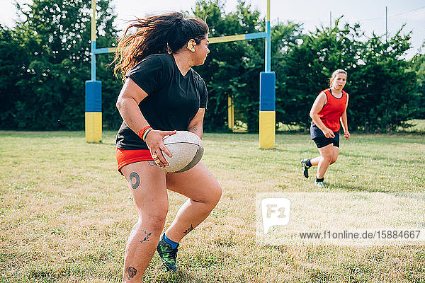 Zwei Frauen rennen auf einem Spielfeld  die eine will der anderen einen Rugbyball zuspielen.