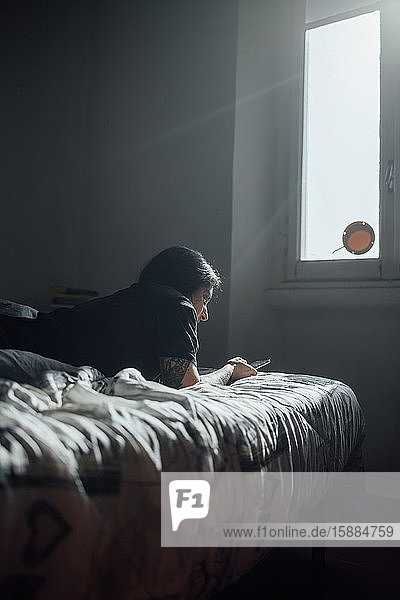 Frau mit tätowiertem Arm liegt auf dem Bett und benutzt während der Corona-Virus-Krise ein Mobiltelefon.