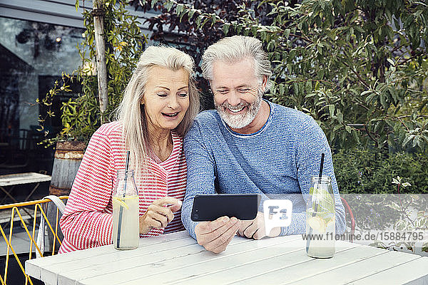 A couple sitting at a table in a garden with drinks in bottles  looking at a mobile phone together.