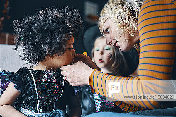 Eine Frau bückt sich  um das Gesicht eines Kindes zu malen  während im Hintergrund ein anderes Kind zuschaut.