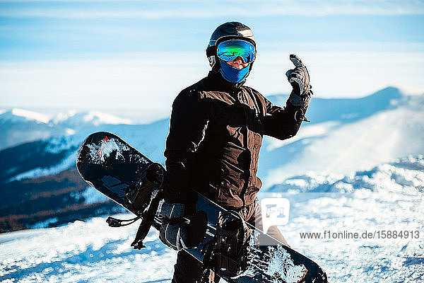 Eine Person  die einen schwarzen Skianzug  einen Helm und eine Schutzbrille trägt und ein Snowboard auf dem Gipfel eines verschneiten Berges hält.
