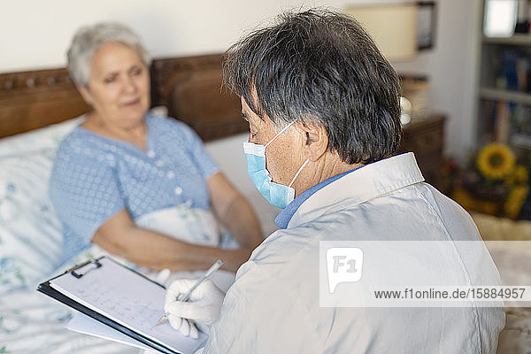 A doctor in a white coat and protective face mask making a home visit to a senior woman patient.