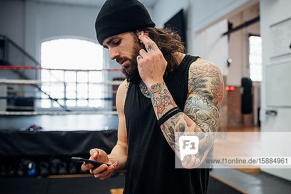 A man standing in a gym wearing a vest and woolly hat with tattooed arms and chest putting a headphone into his ear and looking at a mobile phone.