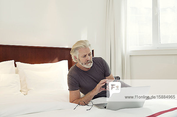 Ein Mann  der auf einem Bett liegt und auf seinen Laptop und sein Mobiltelefon schaut.