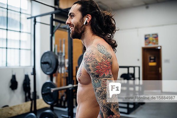 Bare chested man with tattooed arm smiling and standing in a gym.