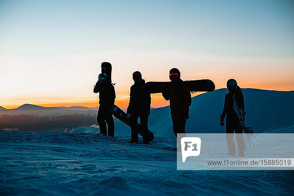 Four people carrying snowboards along a snowy landscape with the sunset in the background.