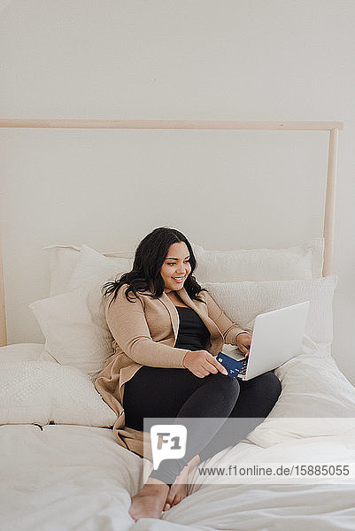 Smiling woman with long dark hair sitting on bed  looking at laptop computer.