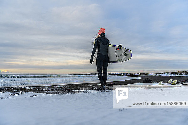 Rückansicht einer Frau  die mit einem Surfbrett in der Hand am Strand steht und aufs Meer hinausschaut.