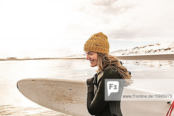 A woman wearing a wetsuit and woolly hat holding a surfboard on a beach with a snowy hill in the background.