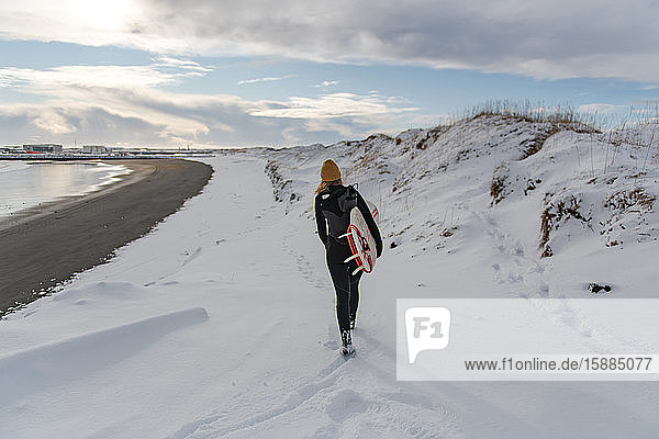 Rear view of a woman wearing a wetsuit and holding a surfboard walking on a snowy beach.