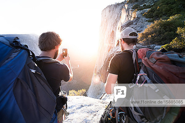 Rückansicht von zwei Männern,  die mit einem Mobiltelefon Fotos von Felsen und einem Sonnenuntergang machen.