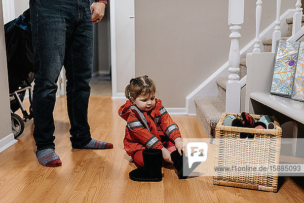A little girl dressed as a firefighter sitting on a wooden floor pulling on black boots