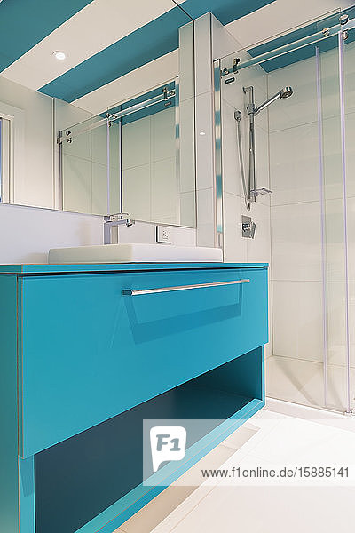 Interior view of modern bathroom with white floor tiles and walls glass shower screen and wall mirror over bright blue vanity unit.