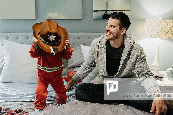 A father sitting on a bed with his child?trying on a cowboy hat.