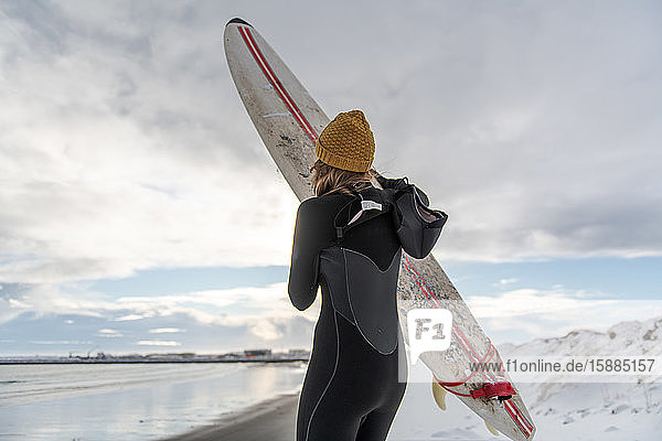 Rear view of a woman wearing a wetsuit and holding a surfboard standing on a snowy beach and looking out to sea.