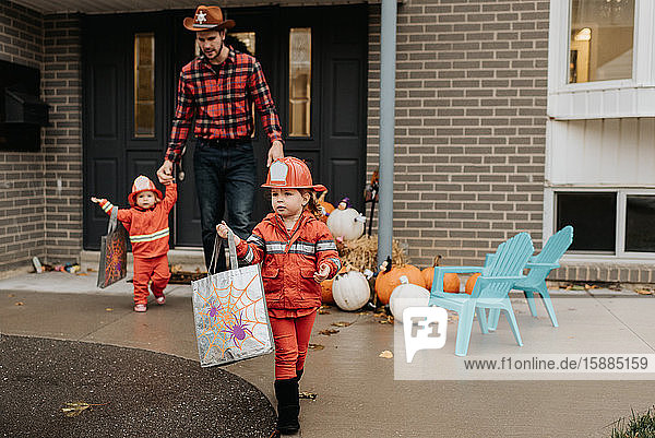 Two children dressed as firefighters walking away from the front door of a house decorated for Halloween with their father.