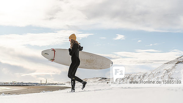 A woman wearing a wetsuit and holding a surfboard walking down a snowy beach and looking out to sea.