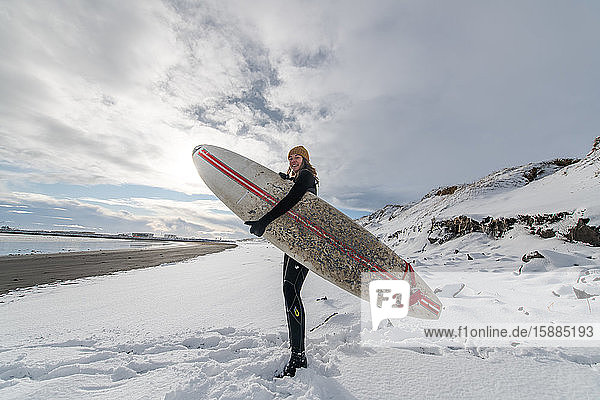 A woman wearing a wetsuit and holding a surfboard standing on a snowy beach and looking out to sea.