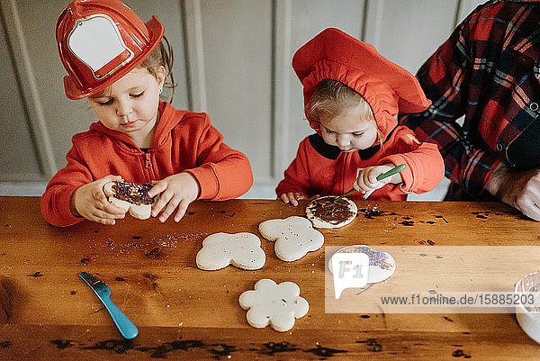 Two children dressed as firefighters at a table decorating cookies with chocolates and sprinkles.