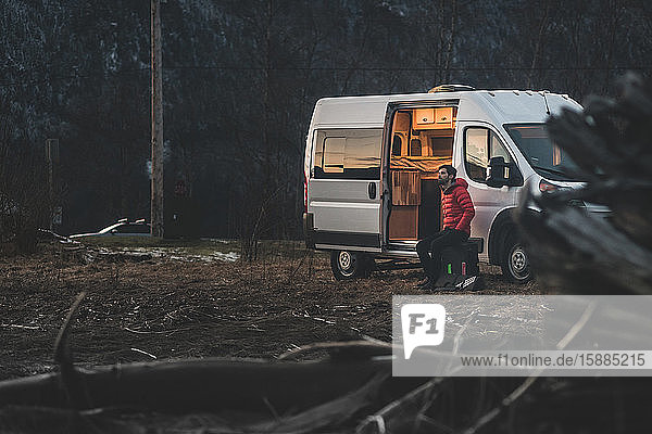 Ein Mann sitzt abends vor seinem Wohnmobil mit dem Wald im Hintergrund.